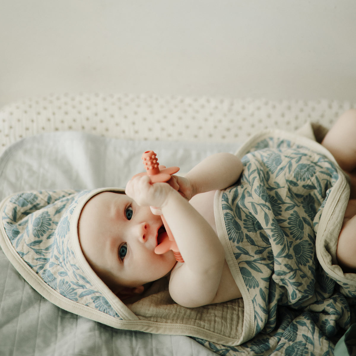 Baby chewing on a coral toy while lying on a floral Mushie Changing Pad Liner.