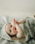 Baby chewing on a coral toy while lying on a floral Mushie Changing Pad Liner.