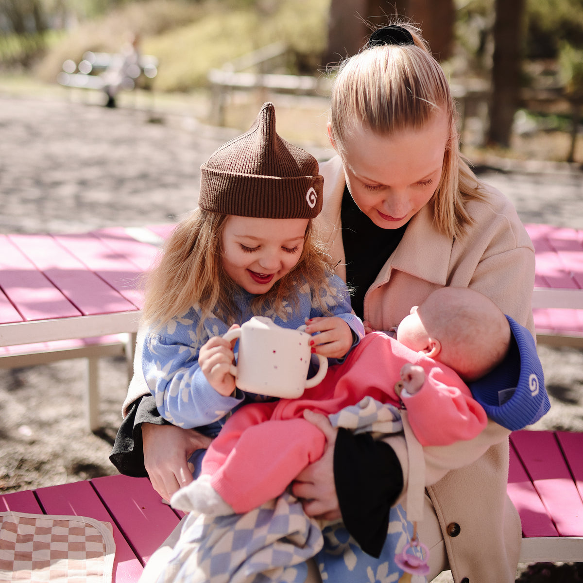 Mom with baby, toddler, and mushie Snack Cup at pink picnic table outdoors.