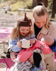 Mom with baby, toddler, and mushie Snack Cup at pink picnic table outdoors.
