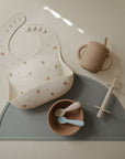 Mushie Silicone Baby Bib set: bib, cup, bowl, and teether on gray mat with adjustable neck strap.