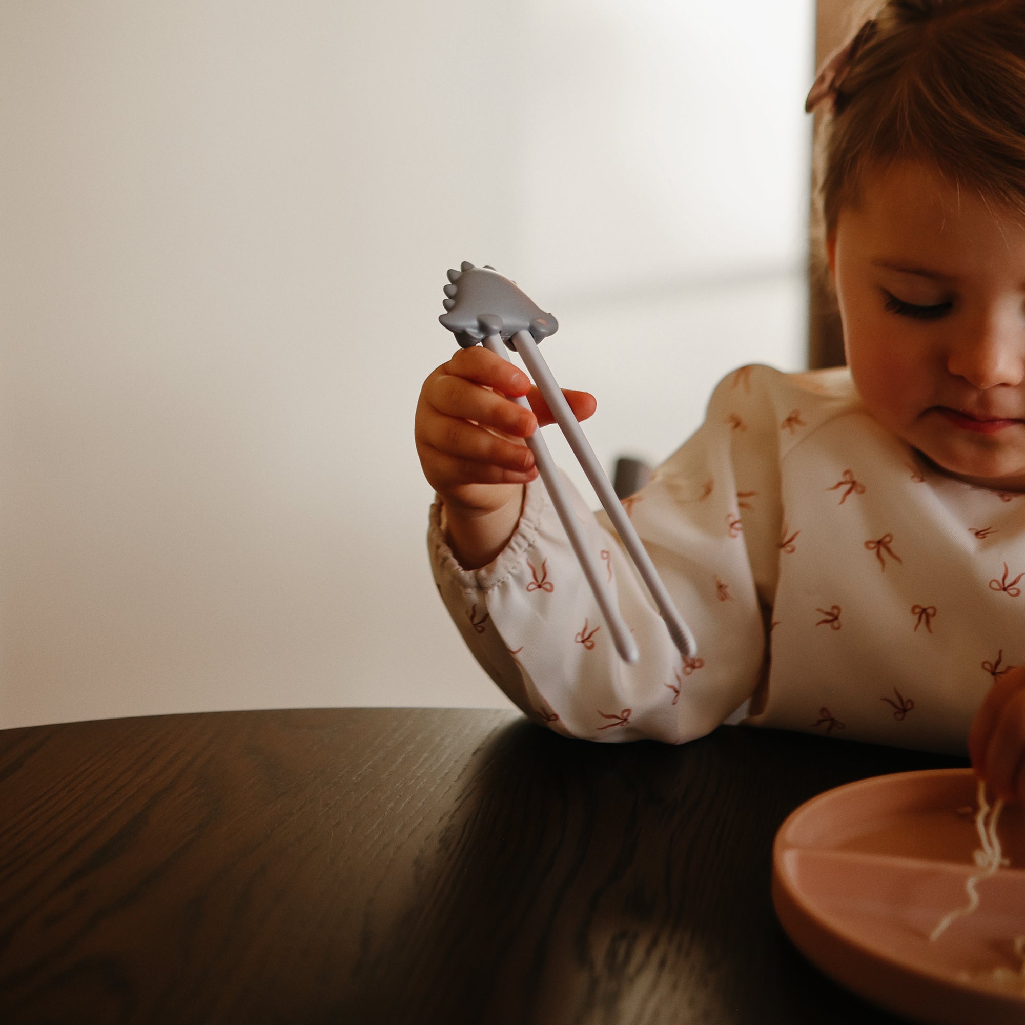 A young child uses mushie Training Chopsticks to pick up noodles from a pink plate.