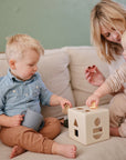 Two toddlers with a mushie Snack Cup play as an adult helps them on a beige couch.