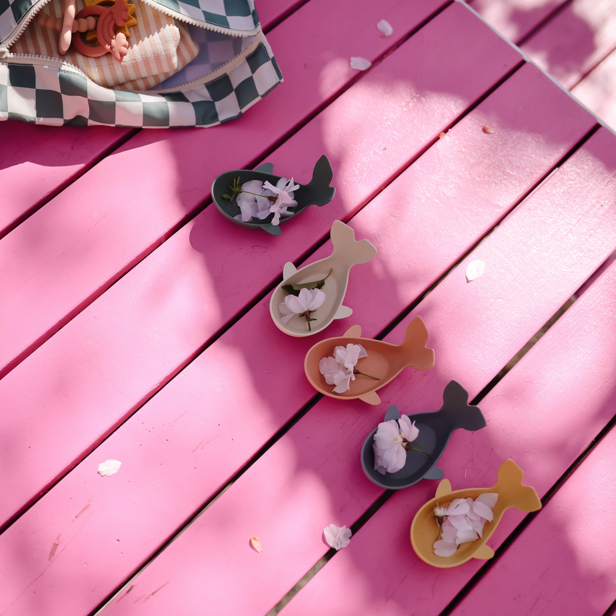 Four Mushie Whale Bath Stackers sit on a pink table under dappled sunlight.