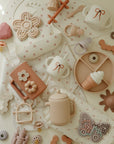 Mushie Snack Cup and pastel toddler toys arranged neatly on a patterned cloth.