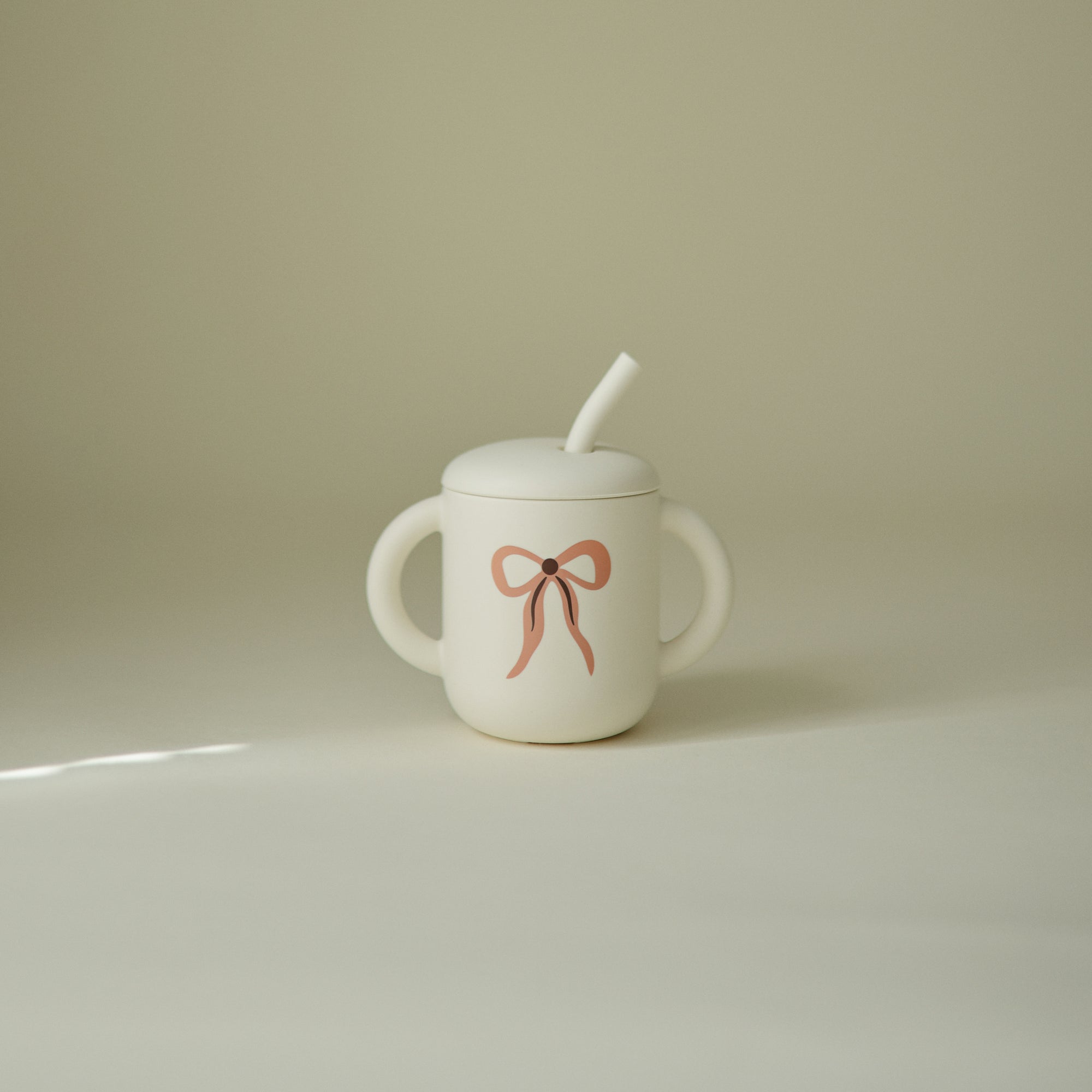 Mushie Silicone Training Cup + Straw: white, two handles, lid, pink bow design.