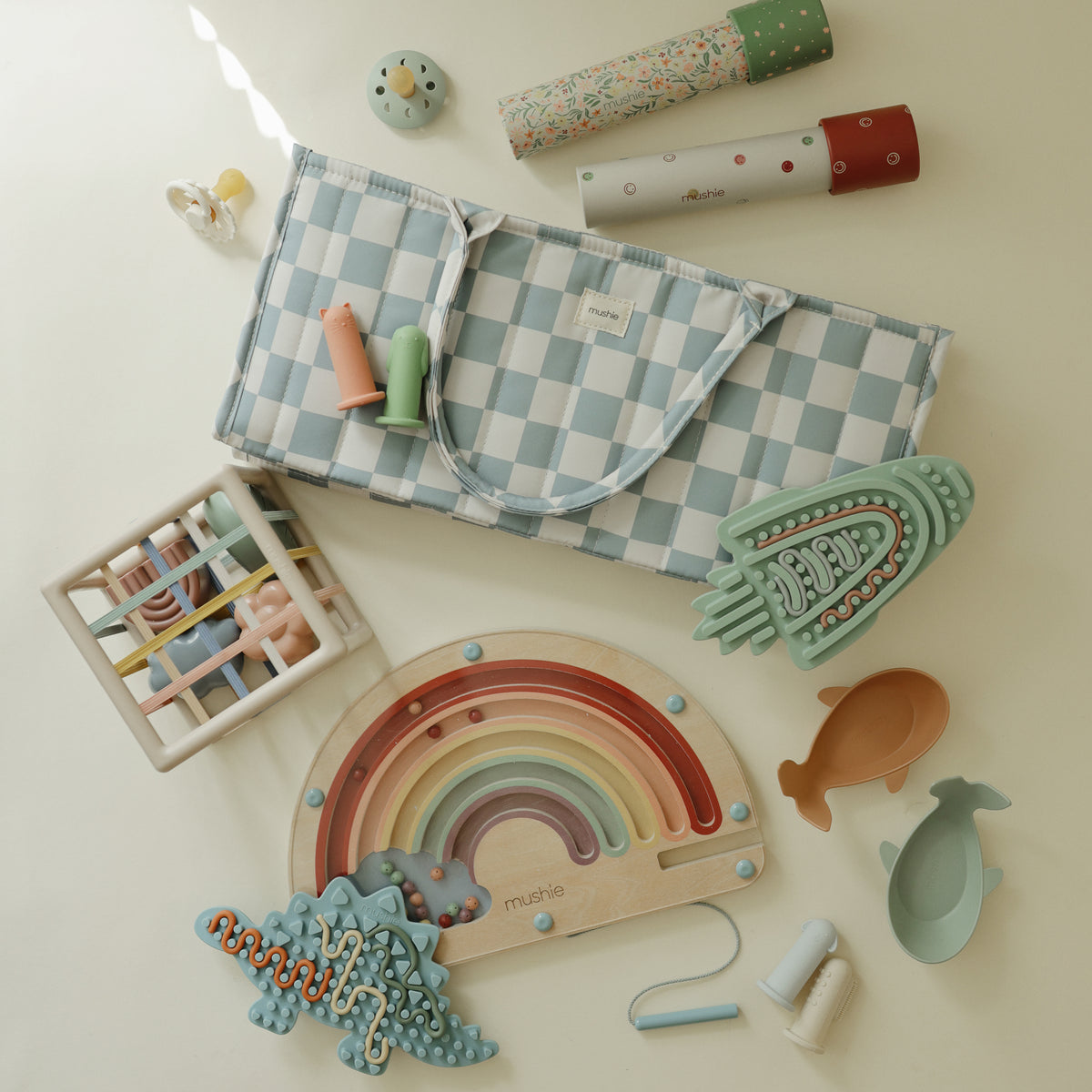Mushie Silicone Sensory Toy with baby toys, a checkered bag, and pacifiers on light surface.