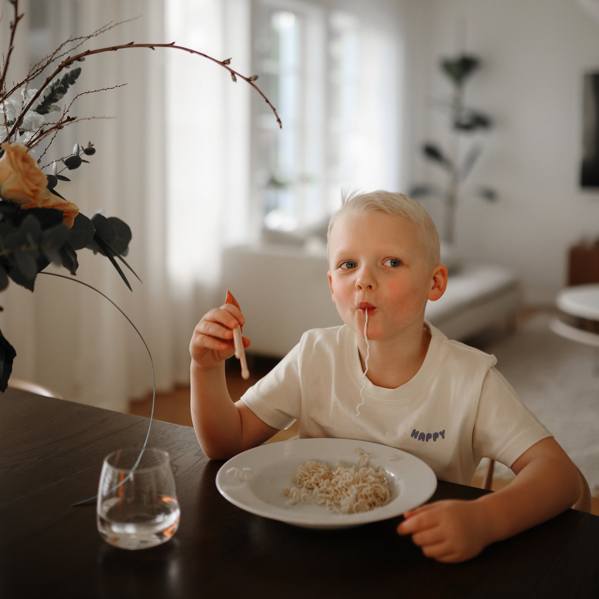 Young boy self-feeding noodles with mushie Training Chopsticks in a modern living room.