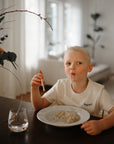 Young boy self-feeding noodles with mushie Training Chopsticks in a modern living room.