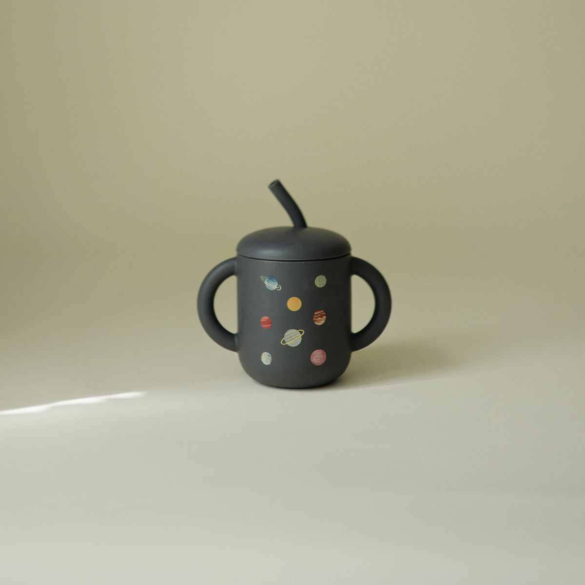 Mushie Silicone Training Cup + Straw in dark color, two handles, planet print, beige background.