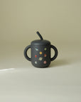 Mushie Silicone Training Cup + Straw in dark color, two handles, planet print, beige background.