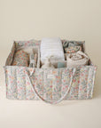 Lifestyle image of the Pastel Blooms Diaper Caddy with folded clothes and baby nursery items on a beige background. 