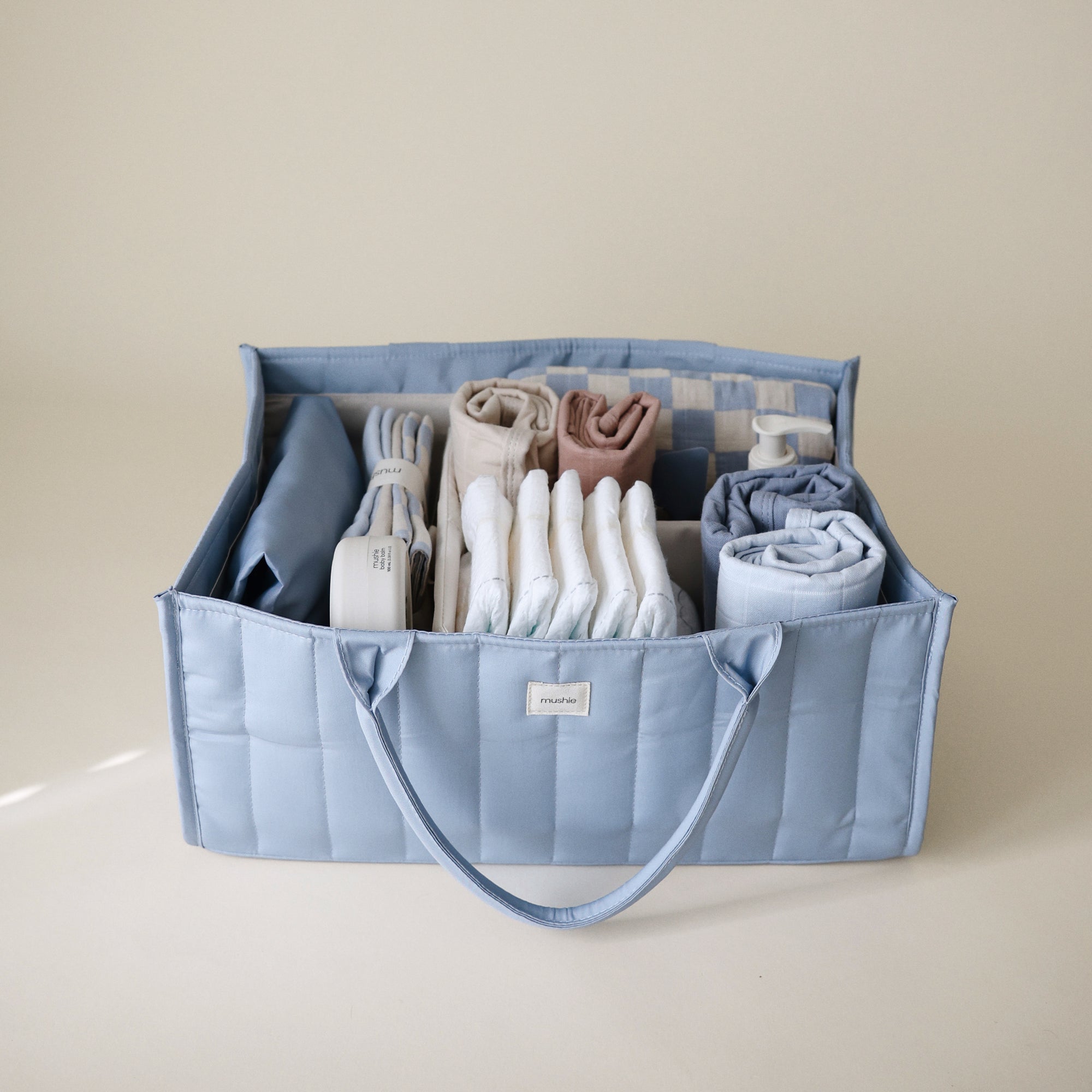 Lifestyle image of the Tradewinds Diaper Caddy with folded clothes and baby nursery items on a beige background. 