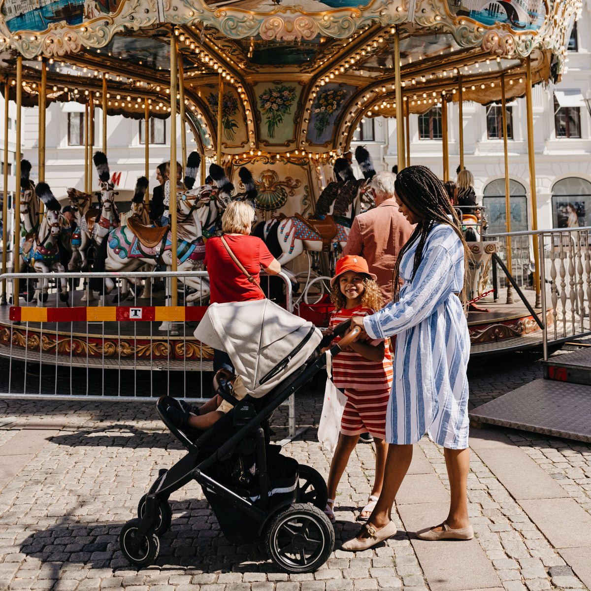 A woman with a mushie Water Resistant Wet Bag and child stand by a stroller near a carousel.