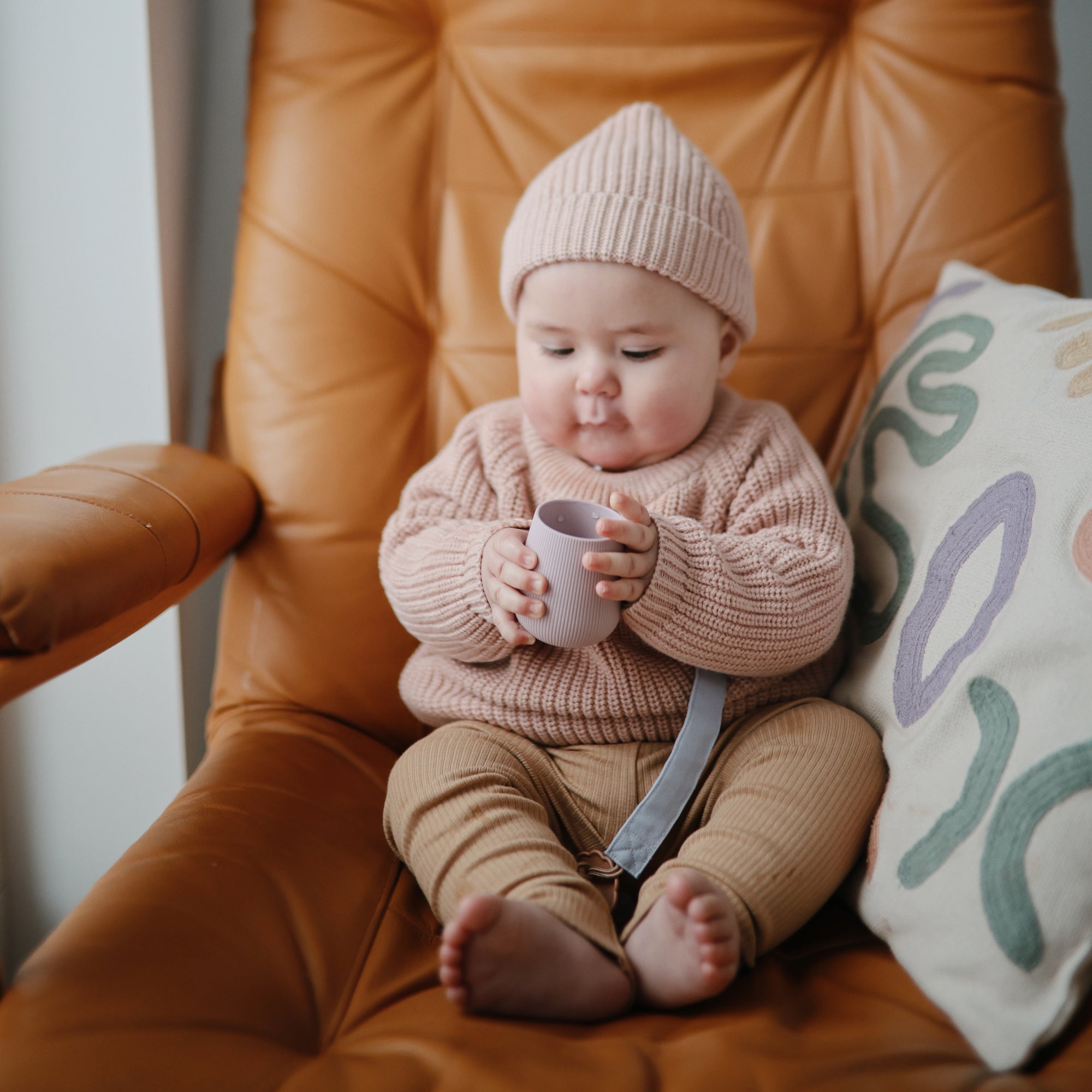 Baby in knit hat holds mushie Linen Pacifier Clip 2-Pack and cup while sitting in a brown chair.