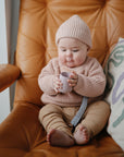 Baby in knit hat holds mushie Linen Pacifier Clip 2-Pack and cup while sitting in a brown chair.