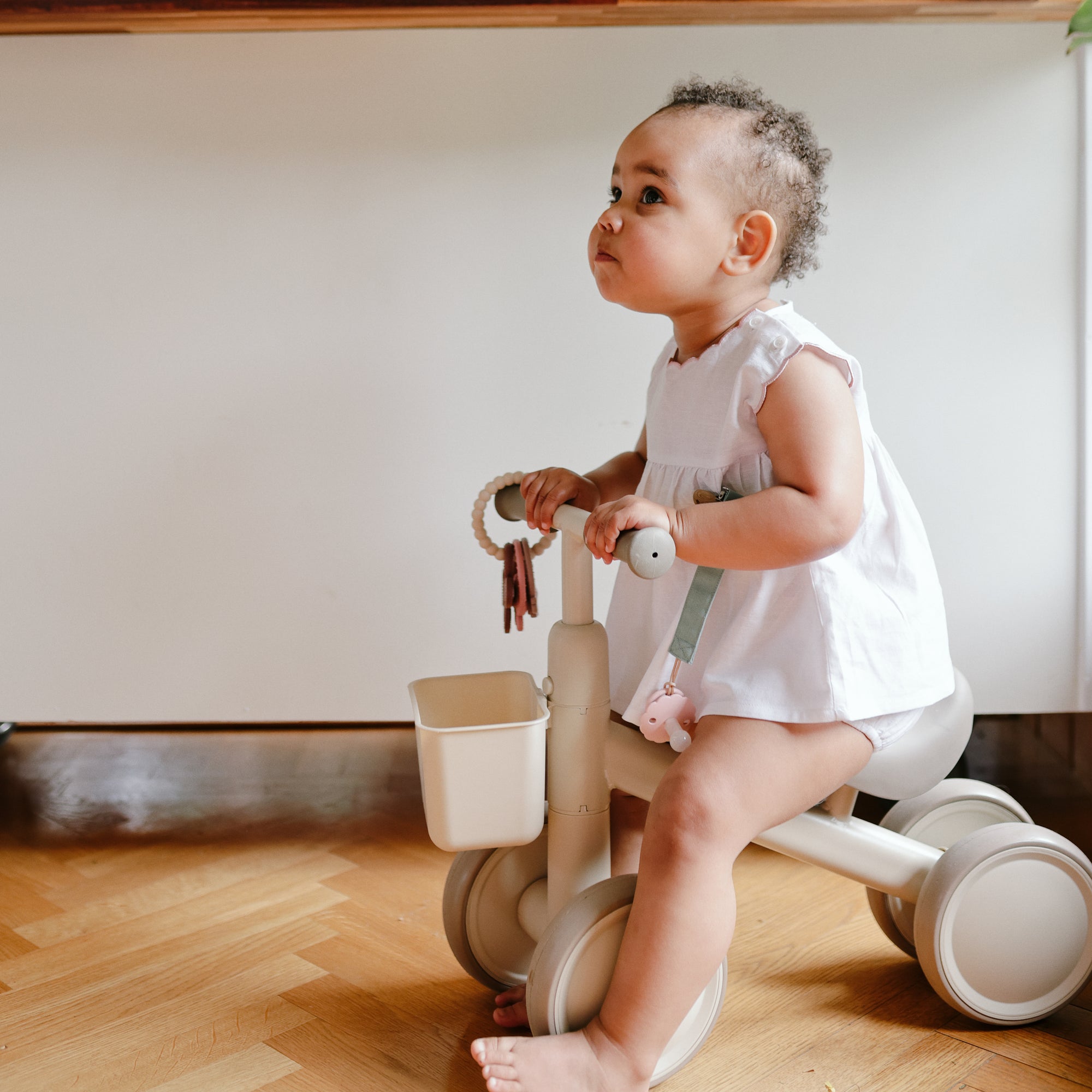 A baby with a mushie Linen Pacifier Clip 2-Pack sits on a tricycle indoors in white clothes.