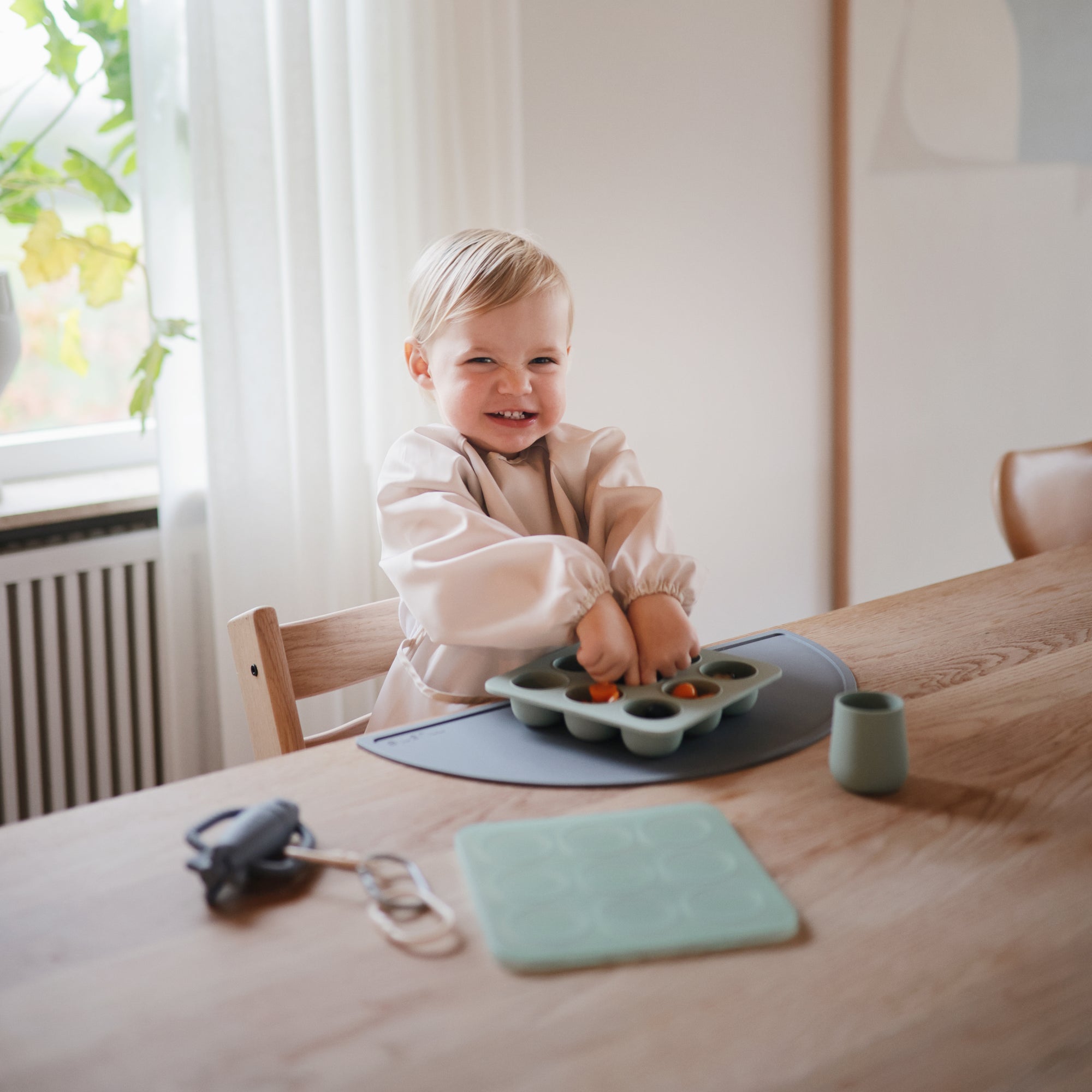 Smiling toddler plays with food in a Mushie Baby Food Freezer Tray at the table.