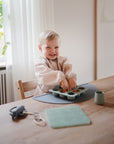 Smiling toddler plays with food in a Mushie Baby Food Freezer Tray at the table.