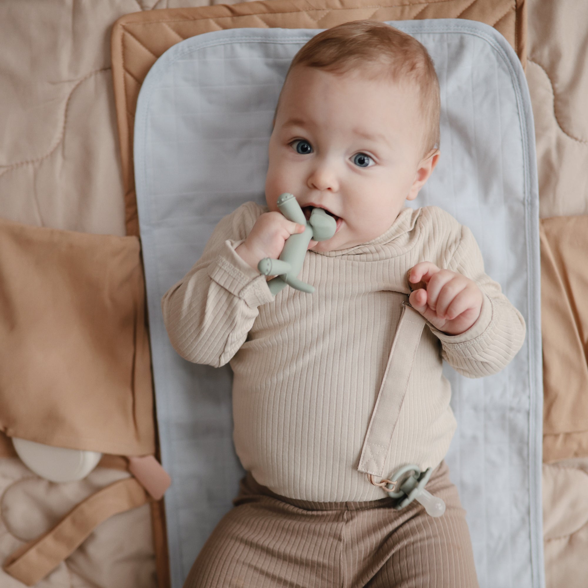 Baby on a mat, wearing beige and chewing on a green mushie Linen Pacifier Clip 2-Pack.