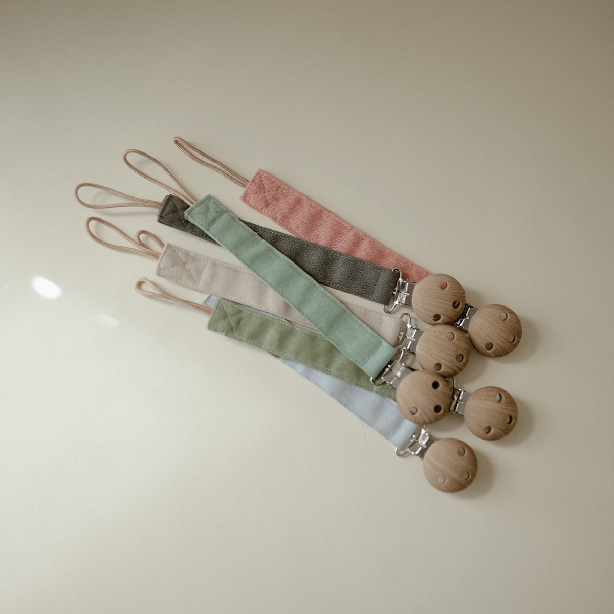 Mushie Linen Pacifier Clip 2-Pack: 4 wooden holders with pastel clips in pink, cream, green, blue.