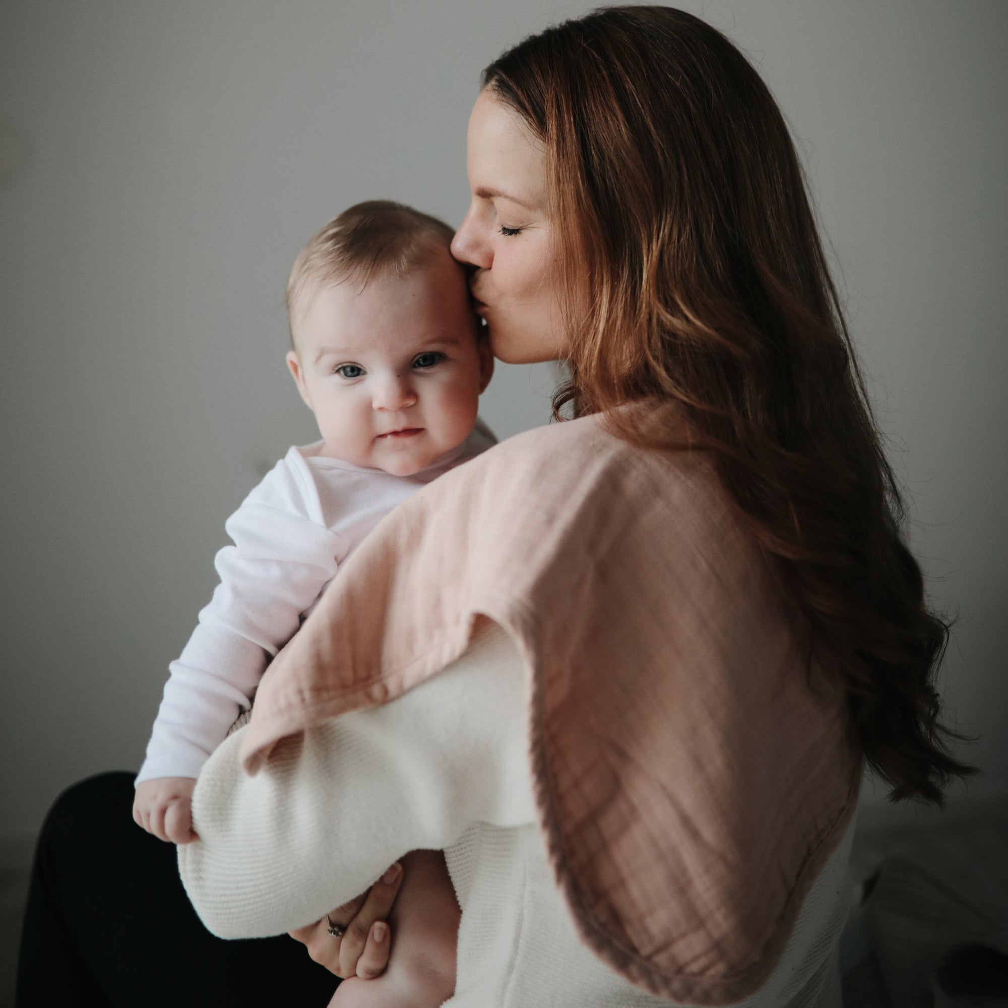 Mother holds and kisses baby with mushie Organic Cotton Muslin Burp Cloth 2-Pack in soft neutrals.