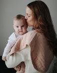 Mother holds and kisses baby with mushie Organic Cotton Muslin Burp Cloth 2-Pack in soft neutrals.