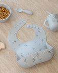 Rylee + Cru Feeding Kit Space: light blue silicone bib by cereal bowl, training cup, and spoon.