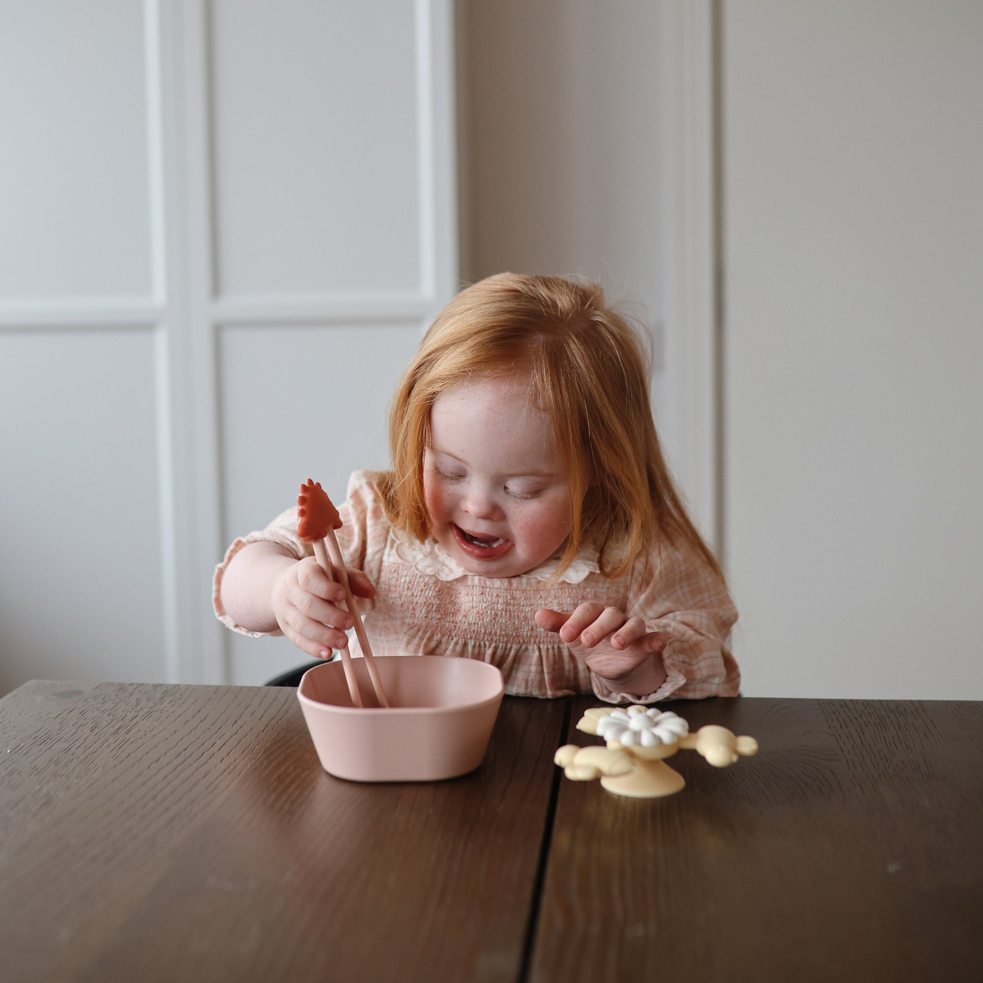 Young girl uses tongs to pick up food from a pink mushie Square Dinnerware Bowl at a table.