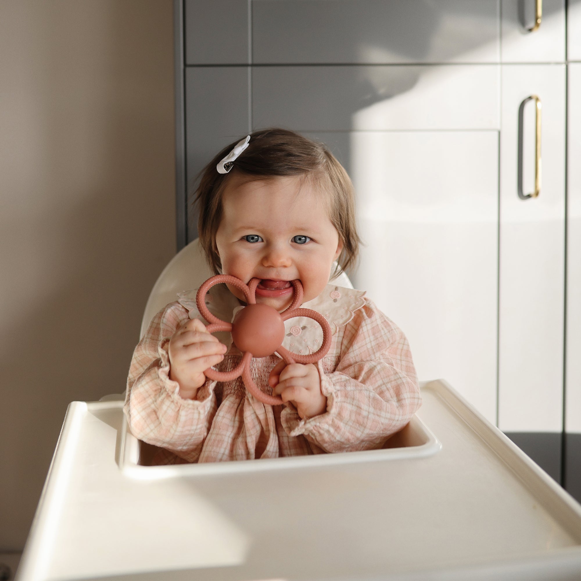 Baby in a high chair smiles, holding a mushie Daisy Rattle Teether.