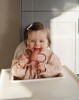 Baby in a high chair smiles, holding a mushie Daisy Rattle Teether.