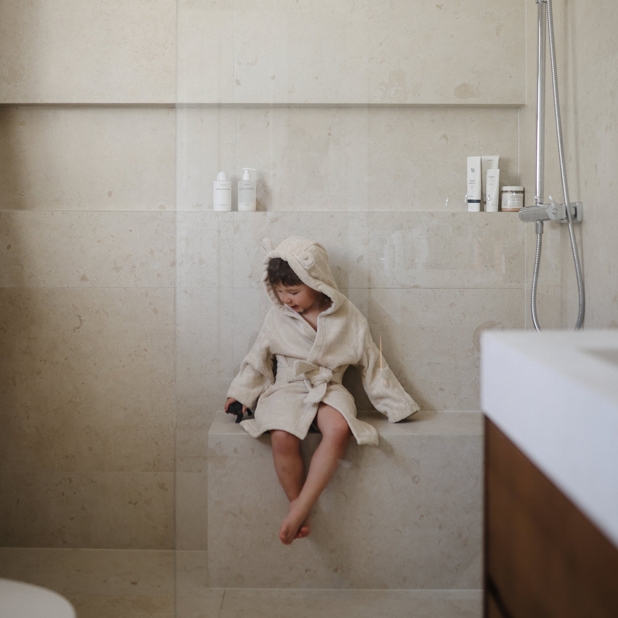 Young child in a mushie Bear Robe sits in a modern, beige bathroom.