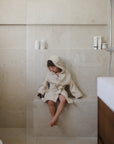 Young child in a mushie Bear Robe sits in a modern, beige bathroom.