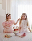 Two girls in pink stand by a birthday cake with a mushie Dinnerware Cutlery Set on the table.