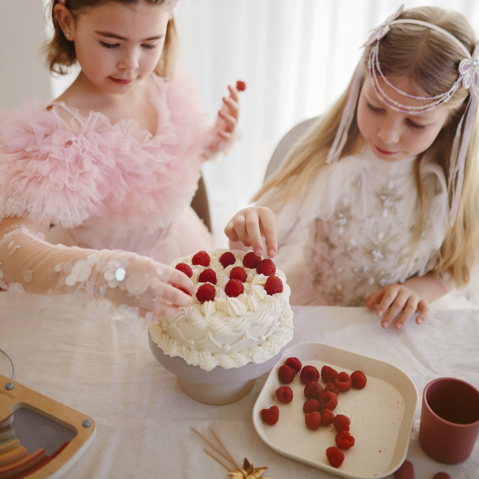 Two girls add raspberries to a cake on mushie Square Dinnerware Plates.