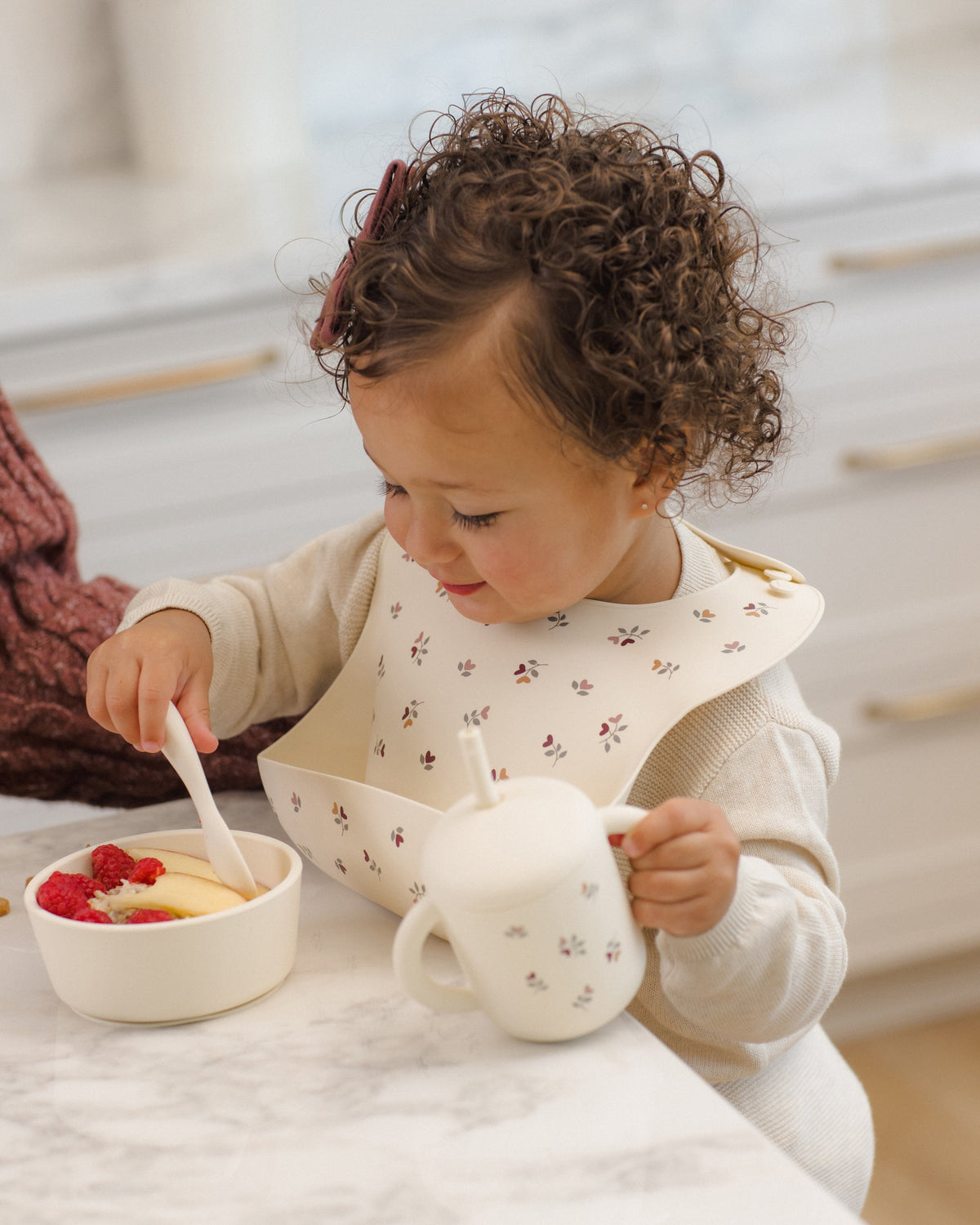 Toddler eats fruit with Rylee + Cru Feeding Kit Heart Ditsy, wearing a silicone bib at counter.