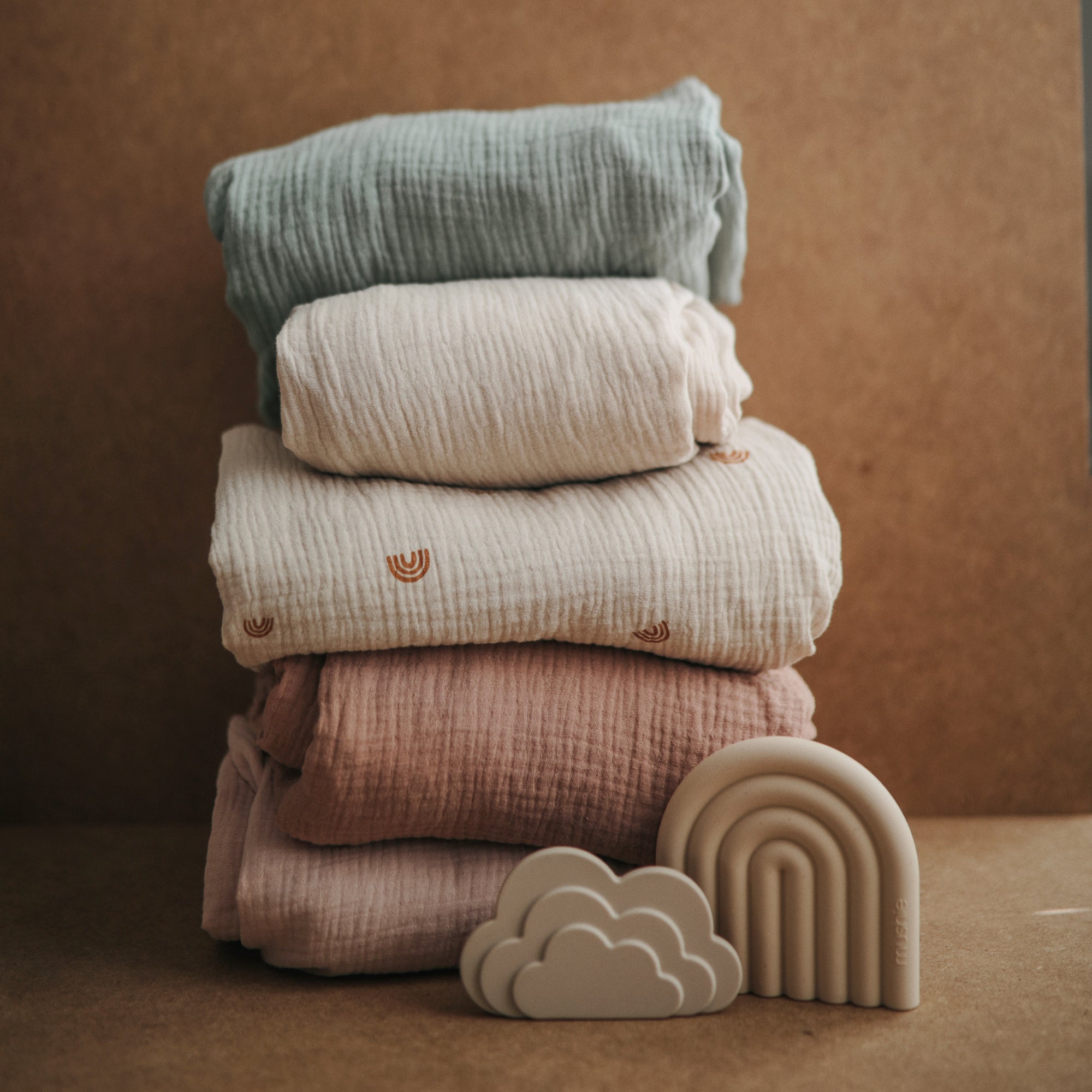 Mushie Extra Soft Muslin Crib Sheets stacked, with cloud and rainbow toys in front.