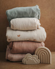 Mushie Extra Soft Muslin Crib Sheets stacked, with cloud and rainbow toys in front.