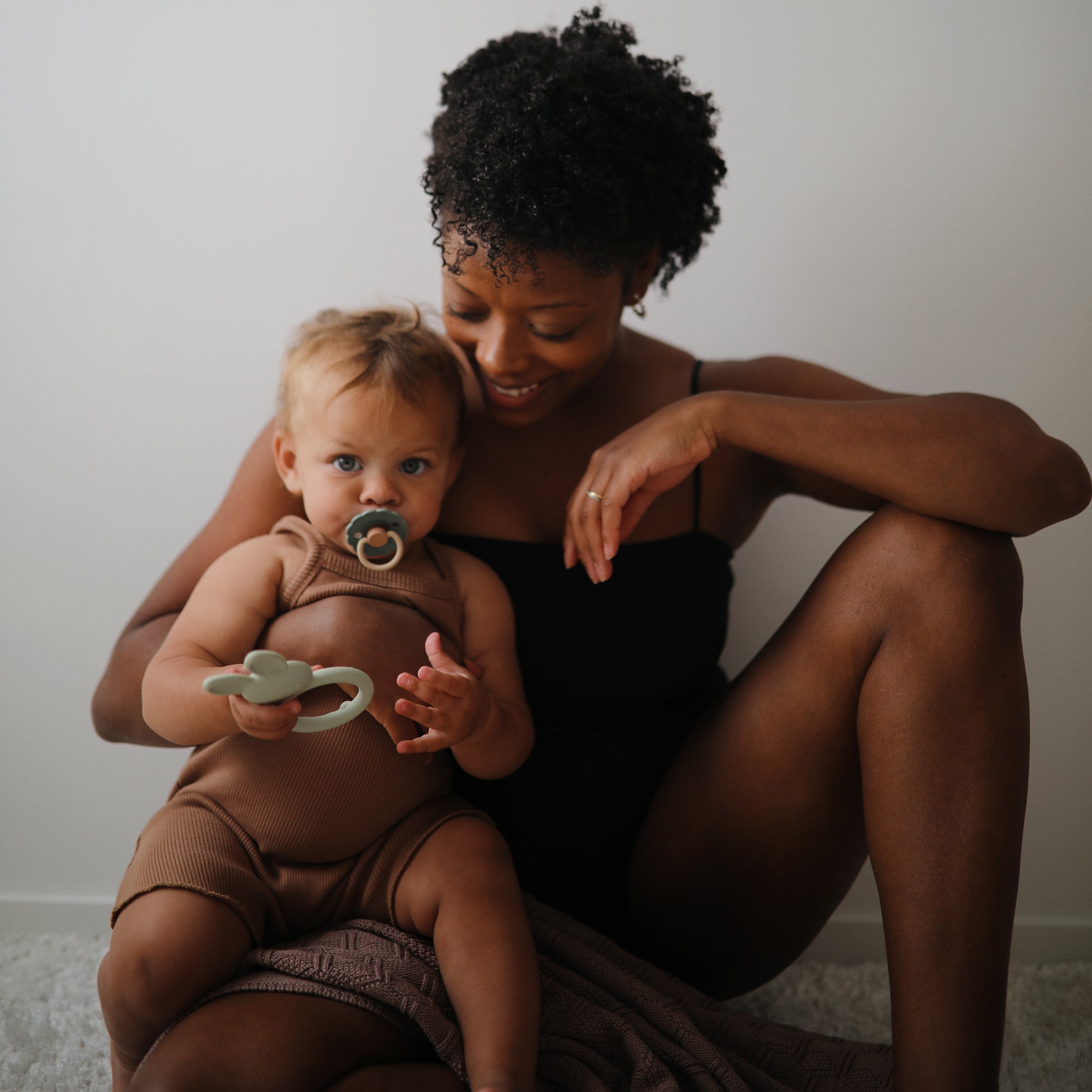 Woman sits holding baby with FRIGG Daisy Natural Rubber Pacifier 2-Pack.