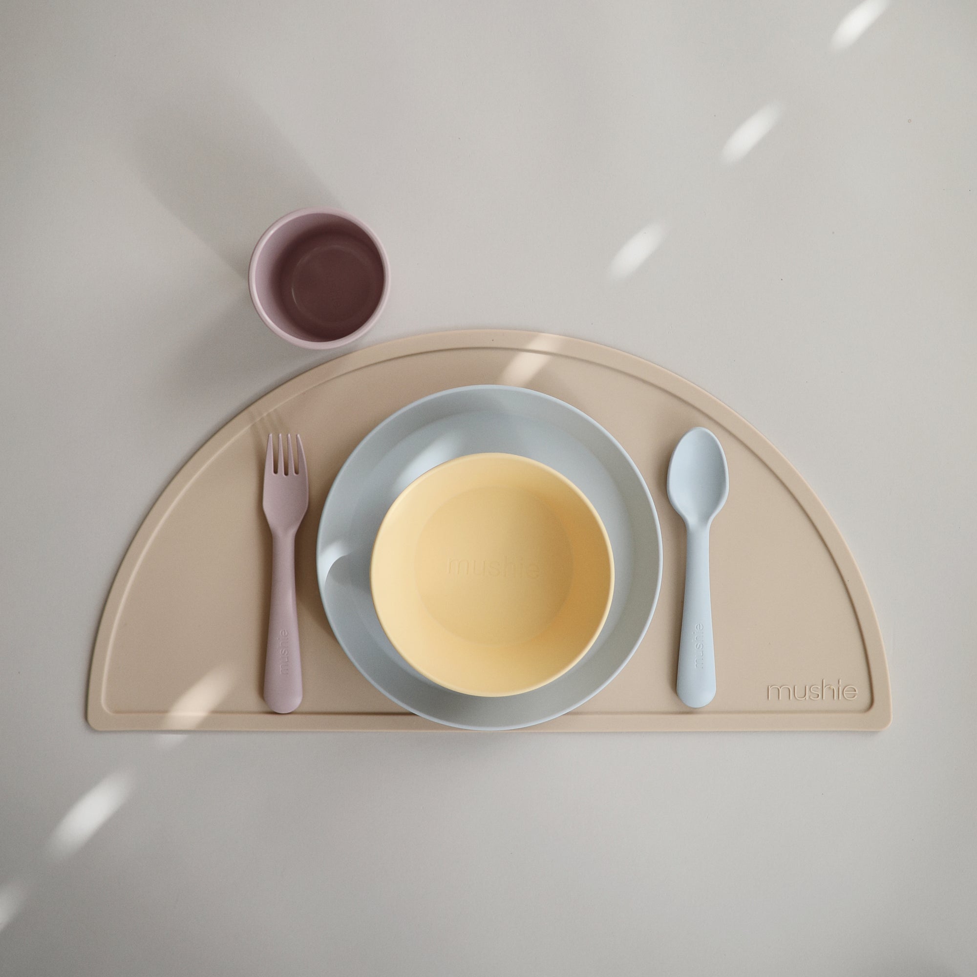 Mushie Dinnerware Fork and Spoon Set: pastel kids’ bowl, plate, cup on placemat.