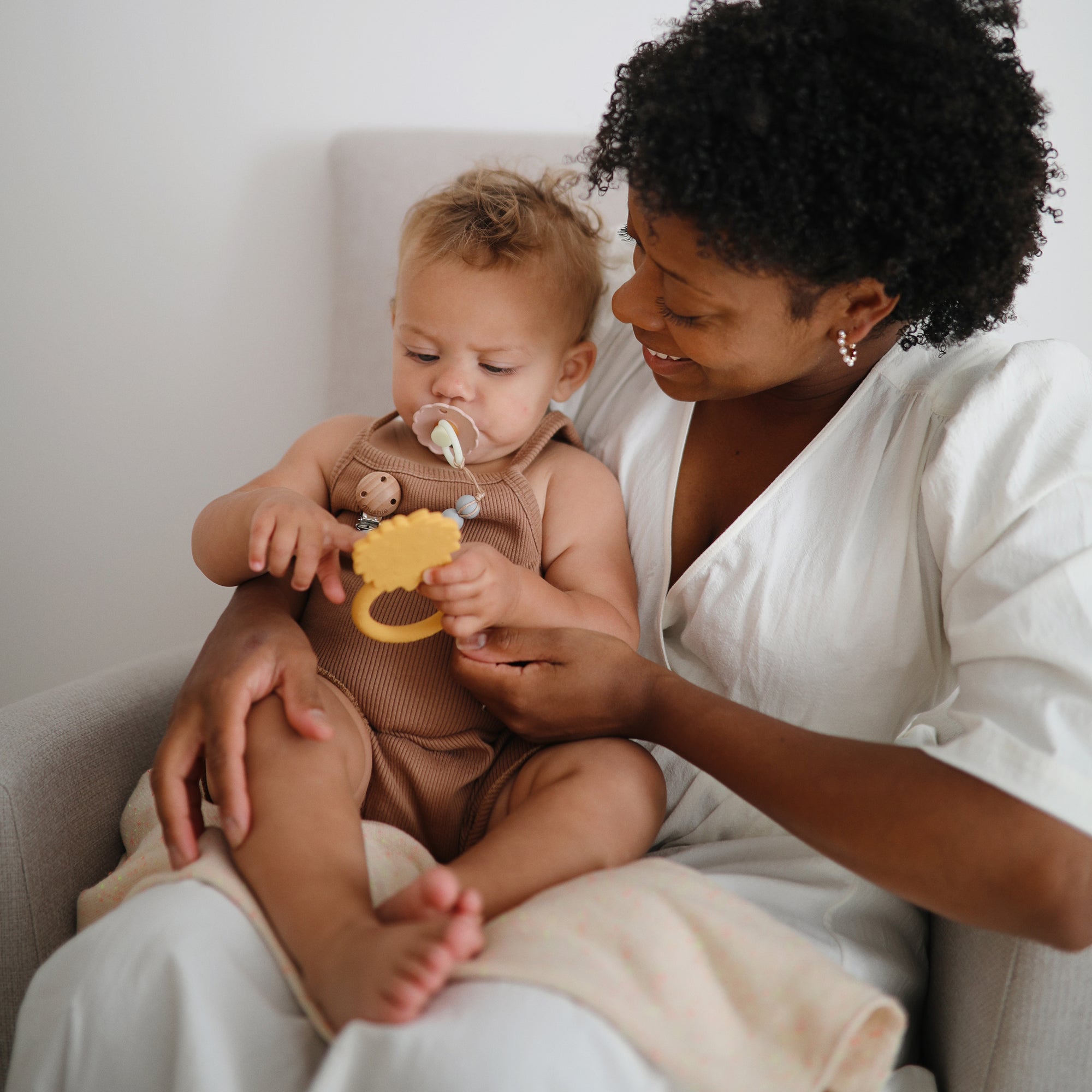 Woman smiles at baby holding Mushie Animal Ring Teether while they sit together on a chair.