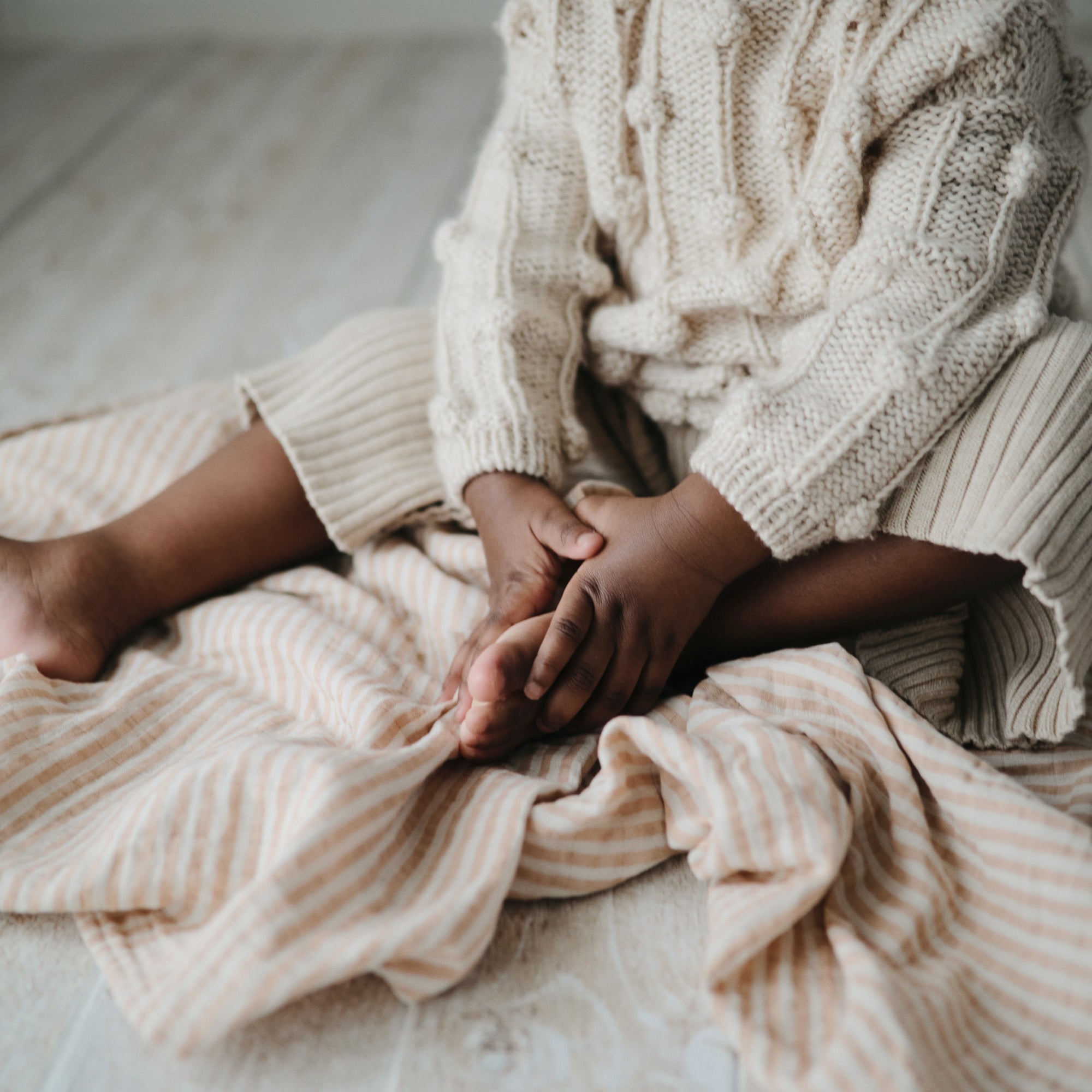 A child sits on a mushie Organic Cotton Muslin Swaddle Blanket, touching their bare foot.