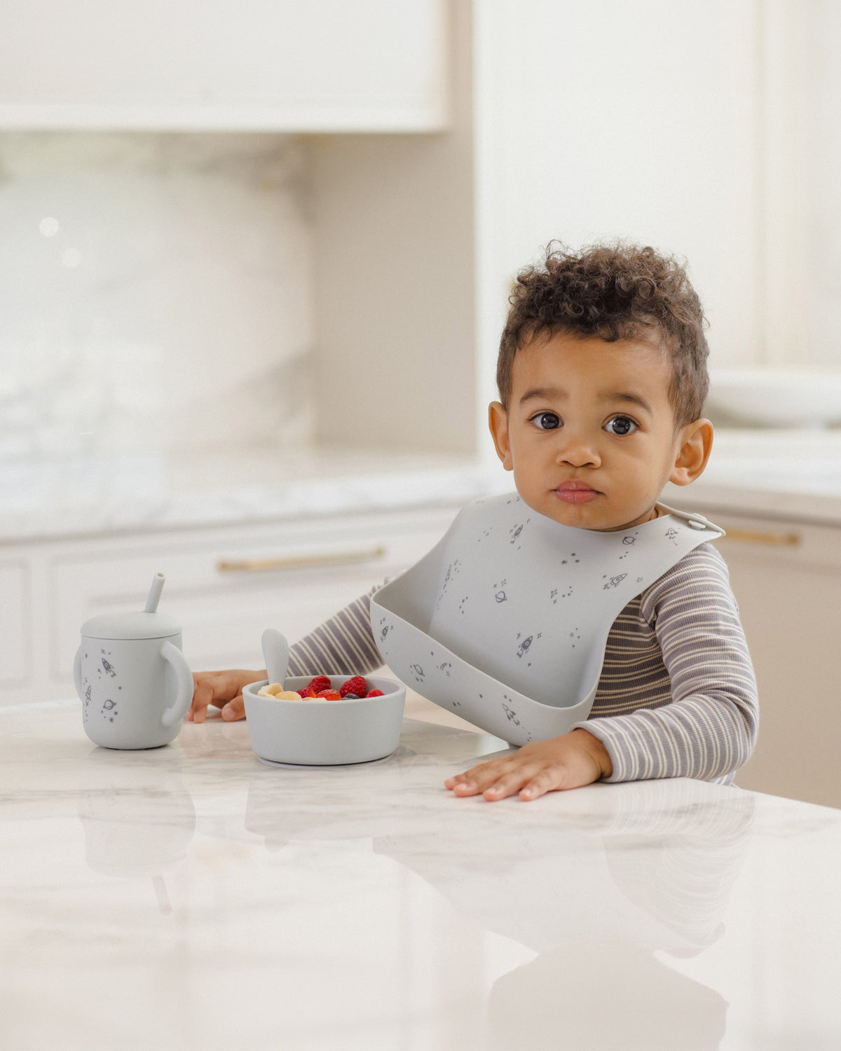 Toddler uses Rylee + Cru Feeding Kit Space: silicone bib, bowl, and training cup at table.