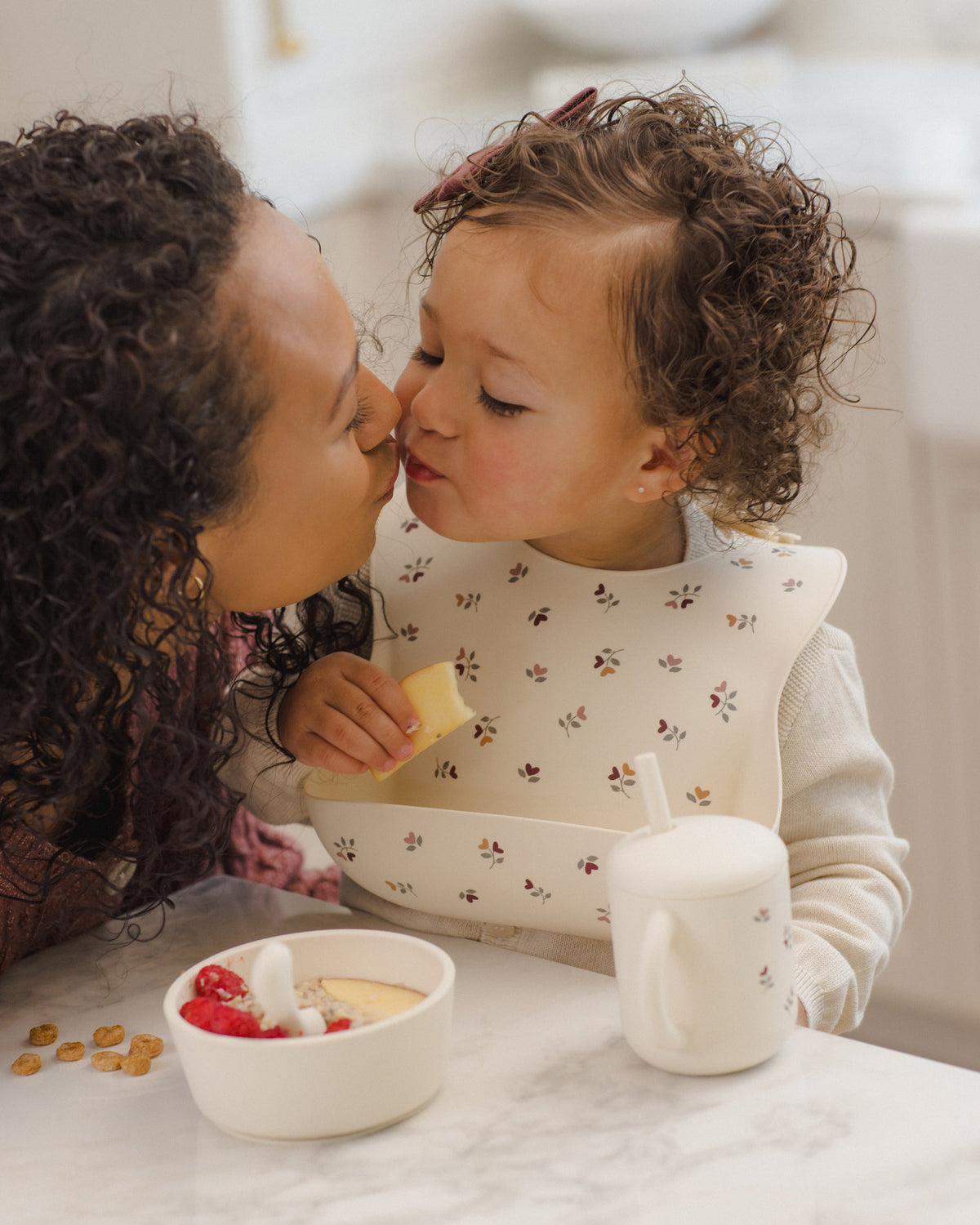 Woman and toddler kiss at table with cereal, berries, sippy cup, and Rylee + Cru Feeding Kit.