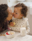 Woman and toddler kiss at table with cereal, berries, sippy cup, and Rylee + Cru Feeding Kit.