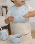 Toddler in a Rylee + Cru Feeding Kit Space bib grabs food near matching bowl and training cup.
