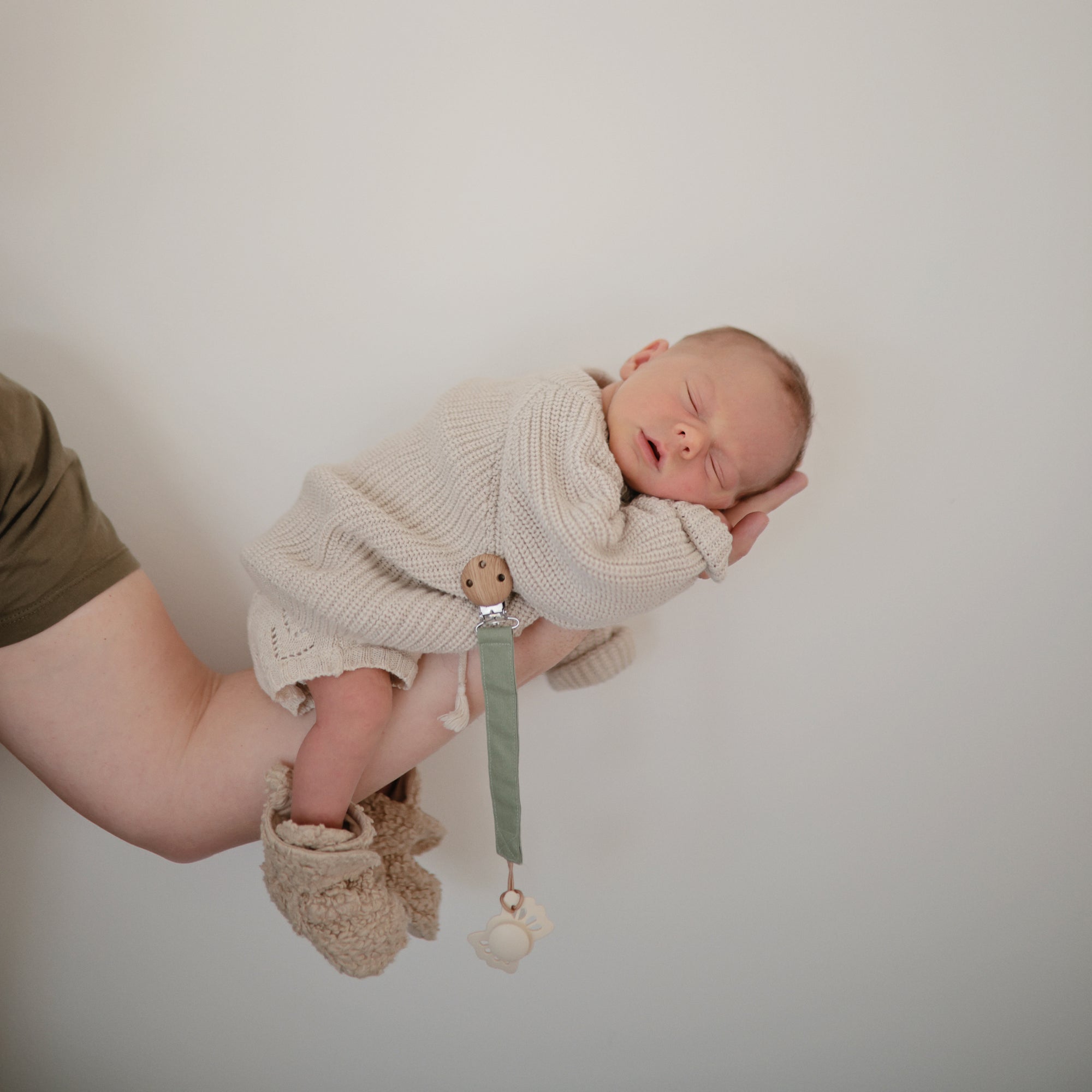 Newborn asleep holding mushie Linen Pacifier Clip 2-Pack against a plain background.