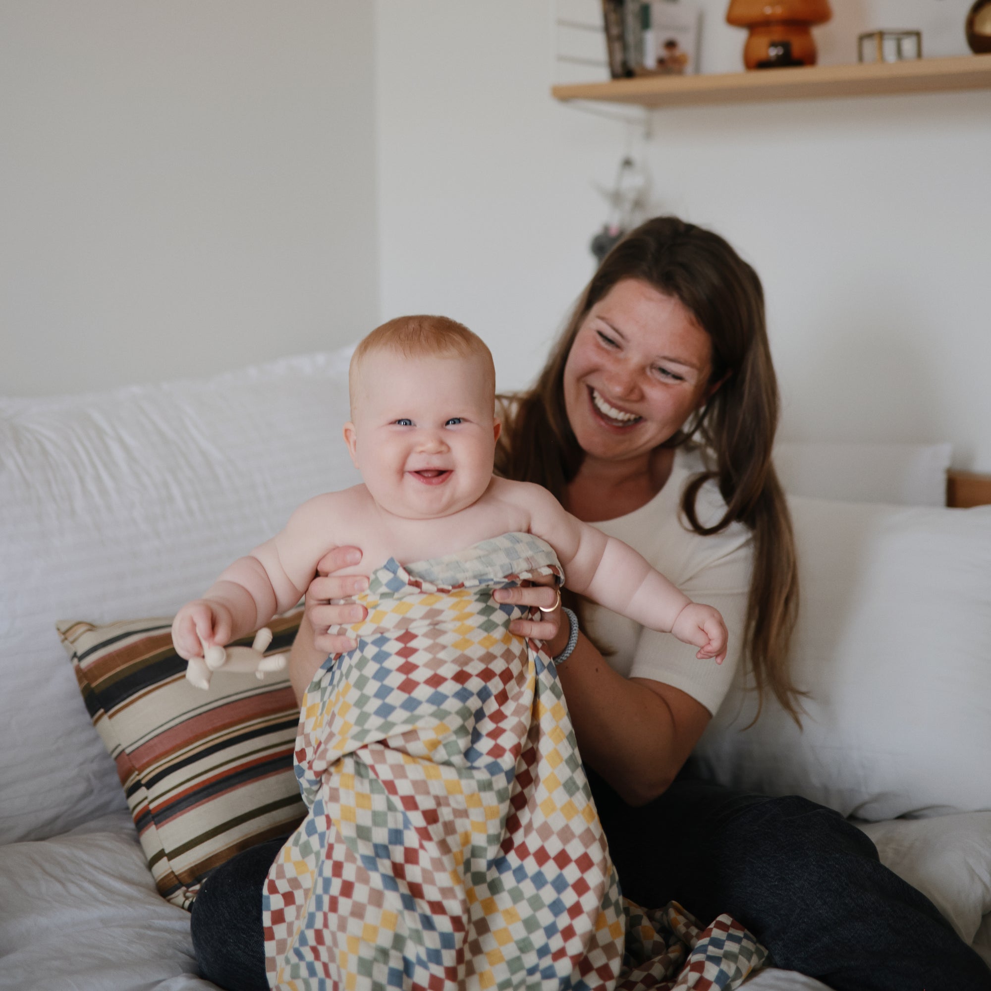 Smiling woman and baby on a bed, baby holding mushie's Figurine Teether and a blanket.