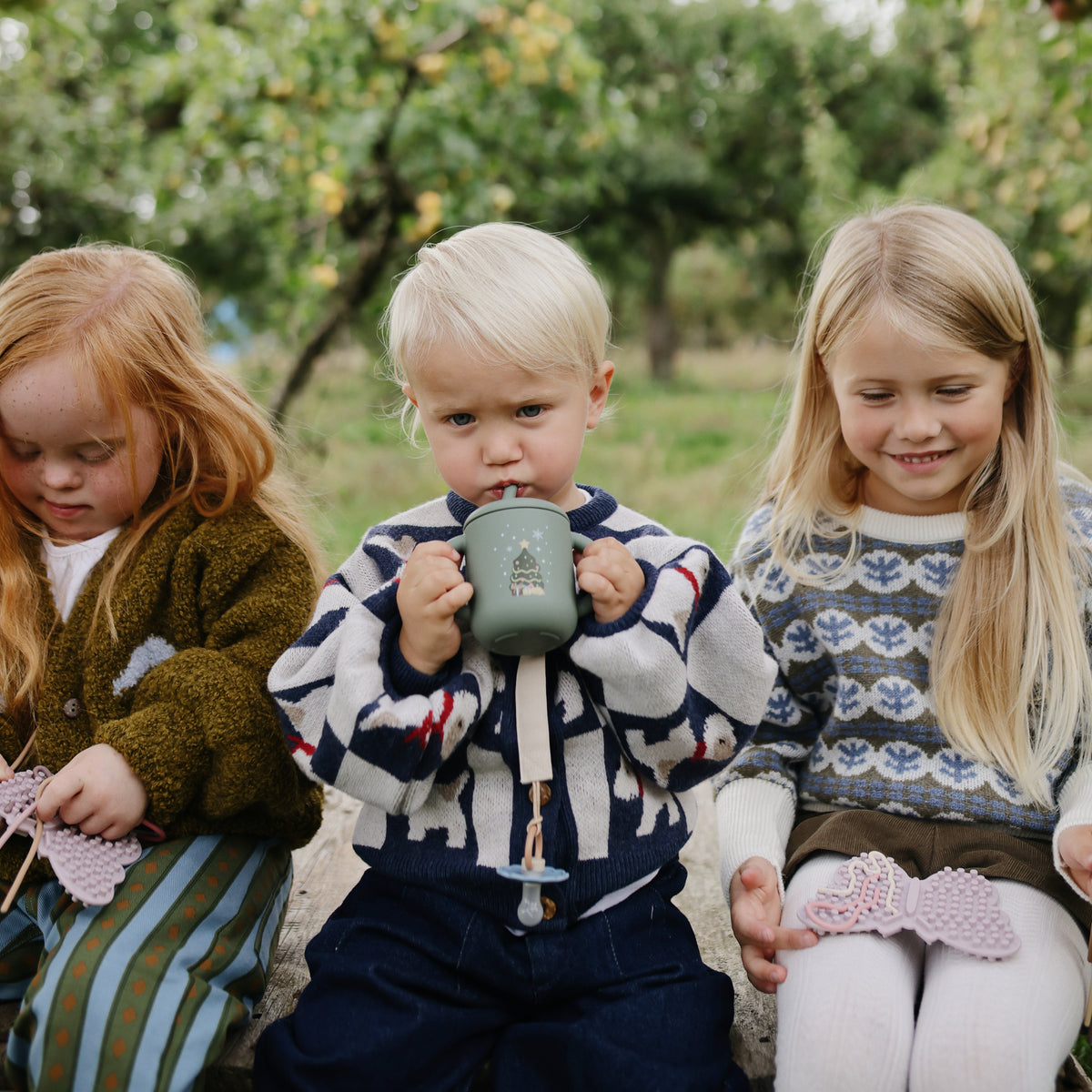 Three kids outdoors: two play with mushie’s Silicone Sensory Toy, one drinks from a mug.