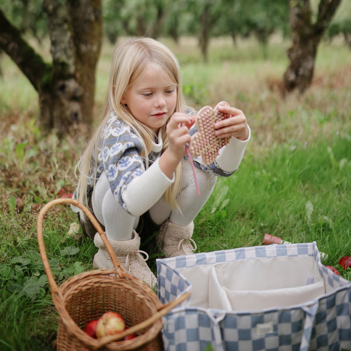 A girl weaves a heart on grass; her mushie Silicone Sensory Toy lies near apples and a basket.